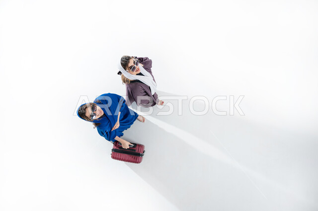 الاستمتاع بإجازة الصيف والهروب من ضغوط الحياة، الاستعداد للسفر الممتع, الاهتمام بالموضة والأزياء العصرية، بورتريه عامودي علوي لسيدتان عربيتان خليجيتان سعوديتان محجبتان ترتديان العباءه الملونه والنظارة الشمسيه تمسك حقيبة الأمتعة بإيماءات السرور والمتعة, خلفيه بيضاء