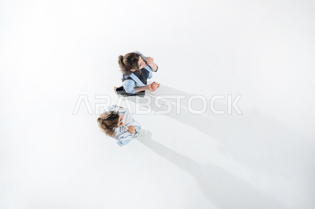 الوقوف بسرور وببشاشة والاستمتاع بالاعتناء بالنفس, مفهوم الاهتمام بالجمال, التسويق لمستحضرات التجميل, بورتريه عامودي علوي لشابتين عربيتين خليجيتين سعوديتين ترتديان الزي الكاجوال الصيفي تقومان بوضع مستحضرات التجميل باستخدام الفرشاه ، مكياج تيتوريال، خلفيه بيضاء