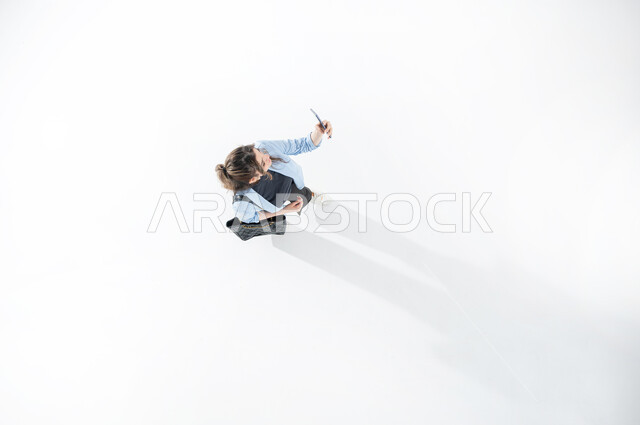 Enjoying taking selfies using mobile phone, vertical top portrait of a Saudi Arabian Gulf woman wearing casual summer dress holding her mobile phone in her hand taking selfies, remembering happy events from mobile phone, white background