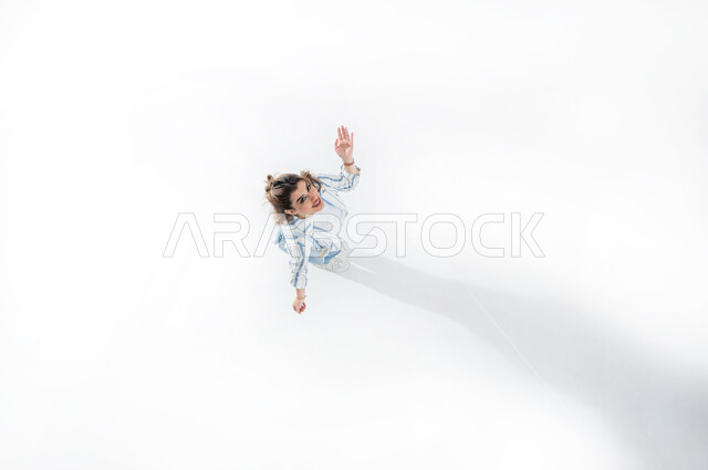 رفع اليدين الى الاعلى للتحيه والسلام، بورتريه عامودي علوي لسيدة عربية خليجية سعودية ترتدي لباس كاجوال صيفي تشير بيدها الى الاعلى بايماءات السعاده الى شئ ما، قضاء العطله الصيفيه مع الاهل والاصدقاء، المناداه باستخدام اليدين، خلفيه بيضاء