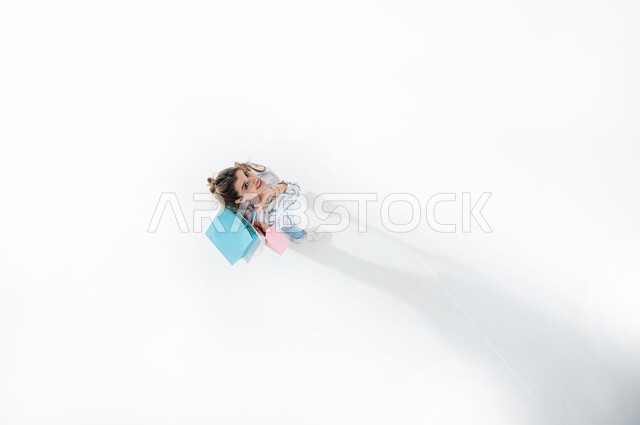 Expressions of happiness with enjoyable shopping and purchasing supplies, taking advantage of seasonal offers and discounts, vertical upper portrait of a young Saudi Arabian Gulf woman wearing casual summer clothes holding colorful shopping bags, shopping in Gulf malls, white background