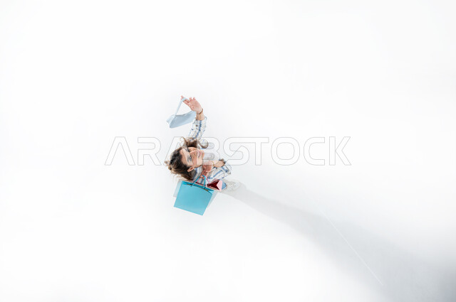 Expressions of happiness with enjoyable shopping and purchasing supplies, taking advantage of seasonal offers and discounts, vertical upper portrait of a young Saudi Arabian Gulf woman wearing casual summer clothes holding colorful shopping bags, shopping in Gulf malls, white background