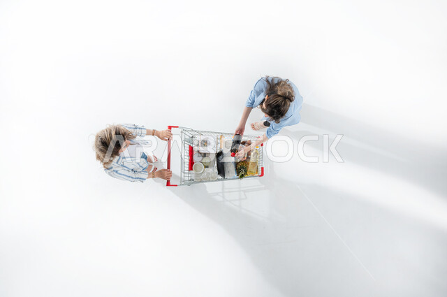 بورتريه عامودي علوي لسيدتان عربيتان خليجيتان سعوديتان تجران بيدهما عربة التسوق ، الاستعداد للتسوق والشراء ، شراء المستلزمات المنزلية ، التمتع بالتخفيضات والعروض ، الاستفادة من الخصومات على المنتجات ، الاستمتاع بالتسوق والترفيه، خلفيه بيضاء