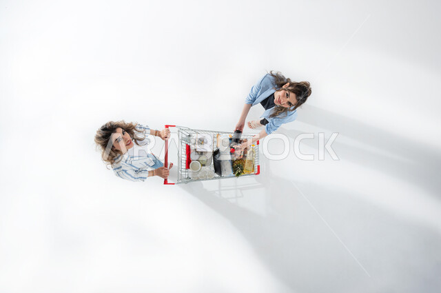 بورتريه عامودي علوي لسيدتان عربيتان خليجيتان سعوديتان تجران بيدهما عربة التسوق ، الاستعداد للتسوق والشراء ، شراء المستلزمات المنزلية ، التمتع بالتخفيضات والعروض ، الاستفادة من الخصومات على المنتجات ، الاستمتاع بالتسوق والترفيه، خلفيه بيضاء