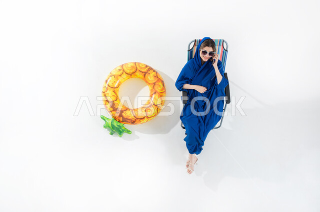 Elegance and interest in appearance, use of modern devices to communicate with family and friends, vertical upper portrait of a veiled Saudi Arabian Gulf woman wearing a colorful abaya and sunglasses sitting on a beach chair making a call, using a mobile phone in free time, white background