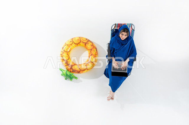 Sitting in public places, using modern devices for chatting and social communication, vertical upper portrait of a veiled Saudi Arabian Gulf woman wearing a colorful abaya sitting on a beach chair working on a laptop, working remotely using a laptop, white background