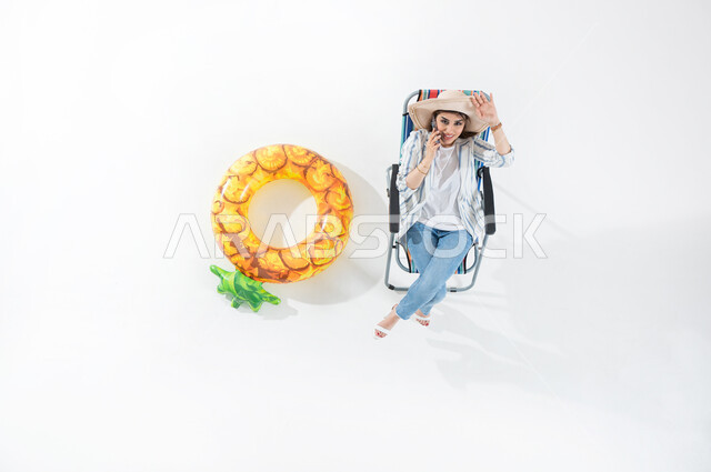 قضاء ايام العطله بتبادل الاحاديث، بورتريه عامودي علوي لشابه عربيه خليجيه سعوديه ترتدي الزي الكاجوال الصيفي تجلس على كرسي الشاطئ تقوم باجراء مكالمه هاتفيه تشير بيدها الى الاعلى بابتسامه ، القاء التحيه والسلام، اوقات ممتعة بتبادل الاخبار والأحاديث، الراحه والاستجمام ، خلفيه بيضاء