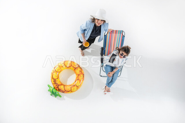 عرض تطبيقات وخدمات السياحة والسفر، الاستعداد للسفر الممتع, الاستمتاع بتناول الفواكه الصيفية المنعشة, بورتريه عامودي علوي لشابه عربيه خليجيه سعوديه ترتدي الزي الكاجوال الصيفي تمسك بيدها الهاتف المحمول بشاشة سوداء فارغة تجلس على كرسي الشاطئ وبجوارها صديقتها تمسك بيدها برتقاله, خلفية بيضاء