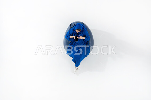 E-marketing and displaying mobile services and applications, using modern technology and techniques, vertical portrait of a Saudi Arabian Gulf woman wearing a colorful hijab and abaya sitting on a comfortable sofa using a mobile phone in her hand, white background