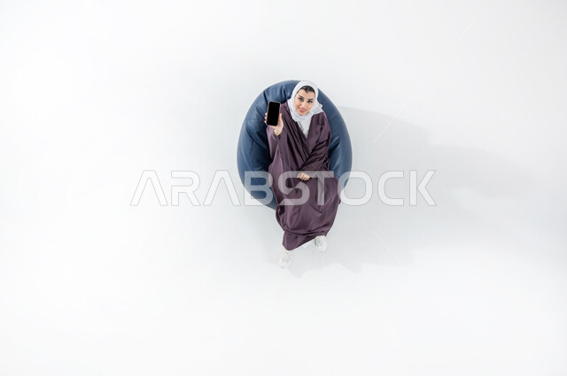 E-marketing and displaying mobile services and applications, using modern technology and techniques, vertical top portrait of a smiling Saudi Arabian Gulf woman wearing a colorful hijab and abaya sitting on a comfortable sofa pointing with a mobile phone with a blank black screen, white background
