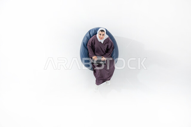 استخدام التكنولوجيا الحديثة، أداء الواجبات الجامعيه وتلقي المحاضرات عن بعد، بورتريه عامودي علوي لسيده عربية خليجية سعودية ترتدي الحجاب والعباءة الملونه تجلس على كنبه مريحه تقوم بالعمل على جهاز الحاسب المحمول تنظر الى الاعلى بابتسامه، خلفية بيضاء
