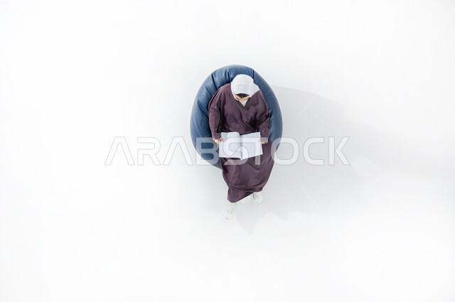 استغلال الوقت في الدراسة والمذاكرة، أهمية التعليم في المجتمع السعودي, بورتريه عامودي علوي لطالبة جامعية عربية خليجية سعودية محجبة ترتدي العباءة وتحمل بيدها كتابا تجلس على كنبه مريحه تقوم بالقراءه, خلفية بيضاء