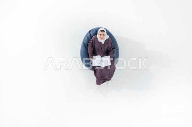 استغلال الوقت في الدراسة والمذاكرة، النظر إلى الكاميرا بإيماءات وجه تدل على السرور، أهمية التعليم في المجتمع السعودي, بورتريه عامودي علوي لطالبة جامعية عربية خليجية سعودية محجبة ترتدي العباءة وتحمل بيدها كتابا, خلفية بيضاء