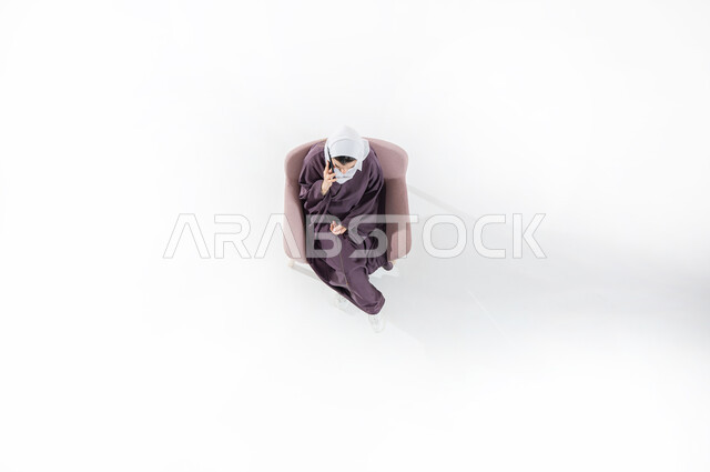 التواصل مع الأهل والأصدقاء، تبادل الأحاديث والأخبار الجيدة، الاتصال الهاتفي عبر الجوال، بورتريه عامودي علوي لشابة عربية خليجية سعودية ترتدي العباءه الملونه والحجاب تجلس على كنبه مريحه من الجلد تقوم بإجراء مكالمة هاتفية عبر الهاتف المحمول، خلفية بيضاء