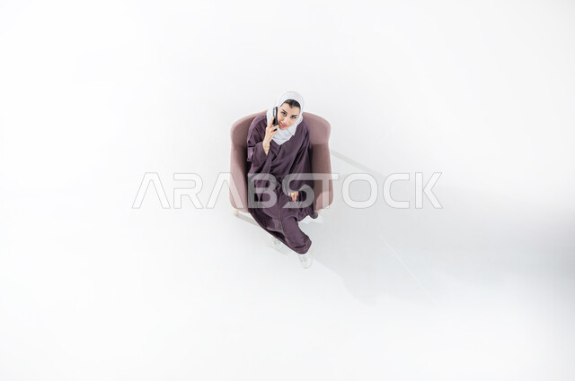 التواصل مع الأهل والأصدقاء، تبادل الأحاديث والأخبار الجيدة، الاتصال الهاتفي عبر الجوال، بورتريه عامودي علوي لشابة عربية خليجية سعودية ترتدي العباءه الملونه والحجاب تجلس على كنبه مريحه من الجلد تقوم بإجراء مكالمة هاتفية عبر الهاتف المحمول، خلفية بيضاء