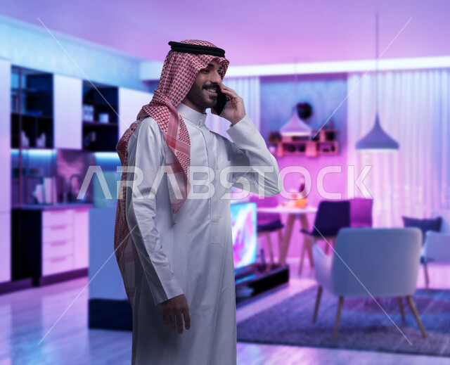 دردشة ومحادثات مع الآخرين، استخدام الهاتف المحمول في الاتصال, رجل عربي خليجي سعودي يرتدي الشماغ والثوب التقليدي يجري مكالمة هاتفية من خلال الجوال، مفهوم صلة الرحم، التواصل مع الأصدقاء والأقارب بإيماءات السعادة، خلفية المنزل