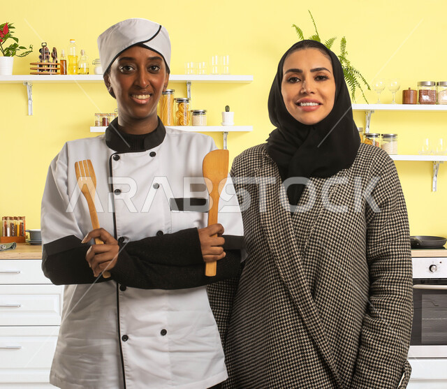 تأهيل وتدريب العاملات المنزليه، شرح طريقة ووصفات الطبخ العربي السعودي التقليدي، صورة مقربة لسيدة عربية خليجية سعودية محجبة وطباخة افريقية تحمل ملاعق خشبية، استقدام الطباخات المرخصة والمؤهلة من الشركات والمكاتب السعوديه، مهن ووظائف نسائية، خلفية المطبخ