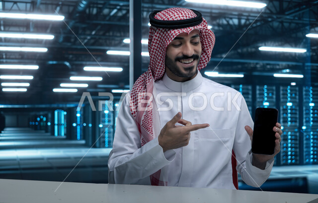 غرفة التحكم بالسيرفرات وخوادم الانترنت، شبكة تخزين البيانات، صورة مقربة لرجل عربي خليجي سعودي مبتسم يرتدي الشماغ والثوب التقليدي يحمل بيده الهاتف المحمول، مركز الداتا سنتر، حظيرة الخوادم والبيانات وتخزينها، التطور الرقمي وتكنولوجيا المستقبل، الاشارة الى شاشة سوداء