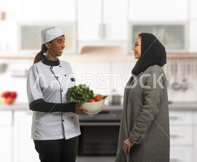 تجهيز مكونات السلطات والاطباق الصحية، صورة مقربة لسيدة عربية خليجية سعودية تقوم بإعطاء تعليمات الطبخ لطباخة افريقية تحمل وعاء خضراوات طازجة، استقدام العاملات و الطباخات المرخصة والمؤهلة من الشركات والمكاتب السعوديه، مهن ووظائف نسائية، خلفية المطبخ