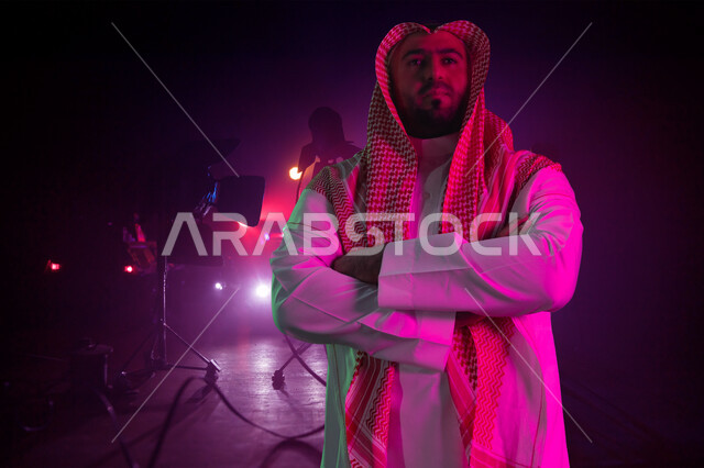 شخصية مهمة سعودية يقف متكتف اليدين في استوديو تصوير، مفهوم الأناقة والرجولة والاهتمام بالمظهر الخارجي، صورة مقربة رجل عربي خليجي سعودي يرتدي الشماغ والثوب التقليدي ينظر إلى الكاميرا بإيماءات الاعتزاز والثقه بالنفس، خلفية اضاءة ملونة  في الاستوديو
