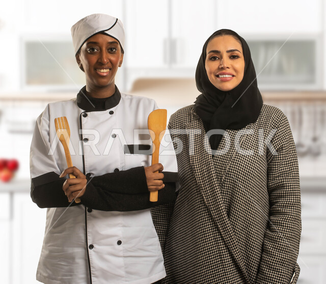 Qualification and training of domestic workers, explanation of the method and recipes of traditional Saudi Arabian cooking, close-up photo of a veiled Saudi Arabian Gulf woman and an African cook holding wooden spoons, recruitment of licensed and qualified cooks from Saudi companies and offices, women's professions and jobs, kitchen background