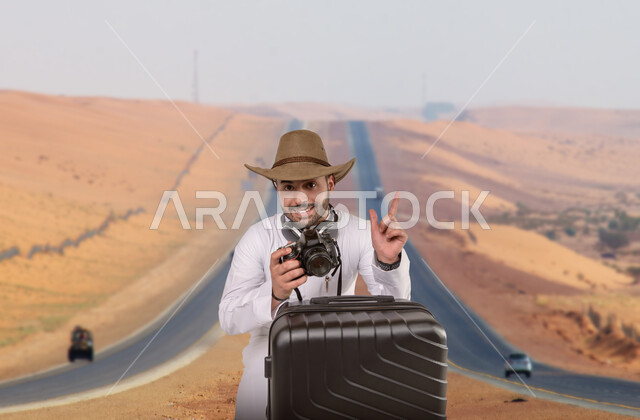 Wild nature in Saudi Arabia, documenting beautiful moments and taking souvenir photos, going around the world for tourism and recreation, spending fun times during the summer vacation, a smiling young Saudi Arabian Gulf man wearing traditional dress and a straw hat standing with a travel bag holding a camera in his hand on a desert road