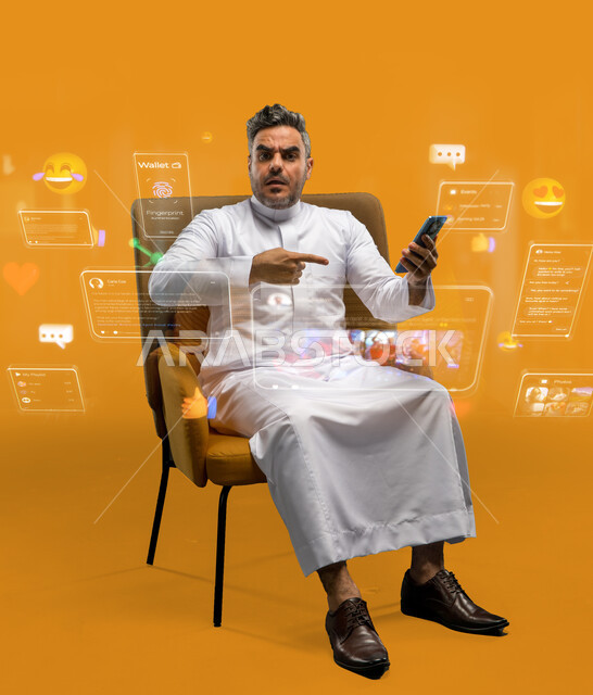 تصفح وسائل التواصل الاجتماعي، استخدام التقنيات الحديثة، رجل عربي خليجي سعودي يرتدي الثوب التقليدي يجلس على كرسي يحمل جوال بتعابير عدم الرضا، خلفيه صفراء مع ايقونات وفقاعات المحادثات والايموجيز، الاشارة إلى الهاتف المحمول بتعابير الغضب والانزعاج
