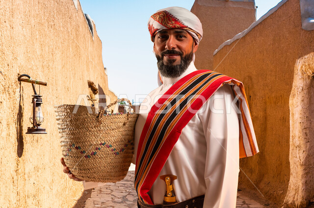 ارتداء ازياء المنطقة الجنوبية, النظر الى الكاميرا بإيماءات الفخر, حرف ومهن يدوية قديمة، ذكرى تأسيس الدولة السعودية 22 فبراير، يوم بدينا 1727 م، رجل عربي خليجي سعودي يرتدي الثوب والغترة مع الشال الجنوبي يمسك بيده سلة مصنوعة من القش، التجول في الاحياء القديمة