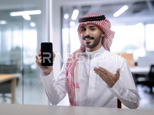 عرض تطبيقات وخدمات الهاتف المحمول, صورة مقربة لشاب عربي خليجي سعودي مبتسم يرتدي الثوب التقليدي والشماغ يحمل بيده جوال بشاشه سوداء فارغة ويشير إليه, النظر إلى الكاميرا بإيماءات السرور, استخدام التكنولوجيا الحديثة للتسوق الالكتروني عبر الانترنت, خلفيه المكتب