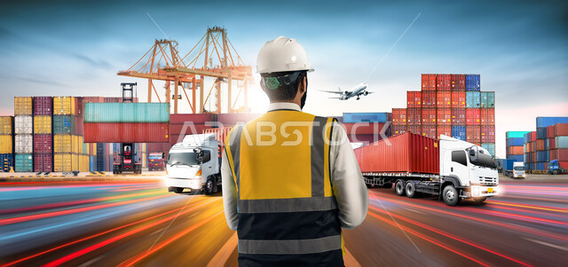 Monitoring and supervising the receipt and transportation of containers, using modern advanced machines in import and export, a picture from the back of a Saudi Arabian Gulf engineer wearing a jacket and a protective helmet standing in a port for loading goods and commercial containers