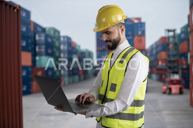 مراجعة قوائم الشحن و الطلبيات، استخدام التقنية و التكنولوجيا و الآلات المتطوره في الإستيراد والتصدير, المراقبة و الاشراف على استلام ونقل الكونتينرات, صورة مقربة لمهندس عربي سعودي خليجي يحمل بيده الحاسب المحمول في ميناء شحن البضائع و الحاويات التجارية