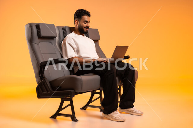 مشاركة تفاصيل الرحلة على مواقع التواصل الاجتماعي، الاستعداد للسفر في إجازة، الجلوس في صالة المطار, بورتريه لرجل عربي خليجي سعودي يرتدي الزي الكاجوال يجلس على المقعد يقوم باستخدام الحاسوب المحمول, أهمية السياحة في السعودية, رحلة عمل جوية، خلفية برتقالية