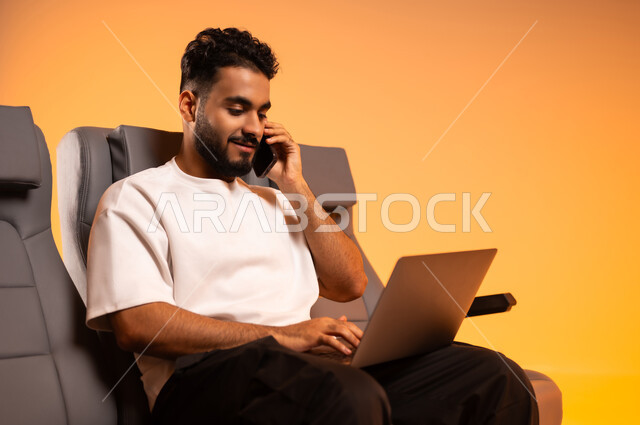 حوارات ودية مع الأهل والأصحاب، التواصل مع الأقارب والأصدقاء عبر جهاز الجوال، الاستعداد للسفر في إجازة، الجلوس في صالة المطار, بورتريه لرجل عربي خليجي سعودي يرتدي الزي الكاجوال يجلس على المقعد يقوم باستخدام الحاسوب المحمول, رحلة عمل جوية، خلفية برتقالية