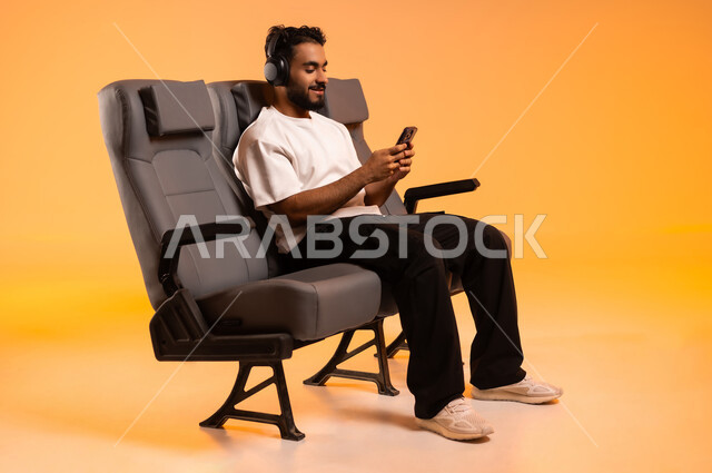 تصفح وسائل التواصل الاجتماعي, الجلوس في صالة المطار، التحدث والدردشة مع الأصدقاء، الاستماع الى الأغاني عبر سماعة الرأس، الاستعداد للسفر في إجازة, بورتريه لرجل عربي خليجي سعودي يرتدي الزي الكاجوال يجلس على المقعد يقوم باستخدام الهاتف المحمول, خلفية برتقالية
