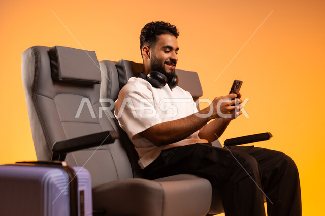 الاستماع الى الأغاني والموسيقى عبر سماعة الرأس، استخدام الأجهزة الحديثة، الاستعداد للسفر في إجازة، الجلوس في صالة المطار, بورتريه لرجل عربي خليجي سعودي يرتدي الزي الكاجوال يجلس على المقعد يقوم باستخدام الهاتف المحمول, أهمية السياحة في السعودية, خلفية برتقالية