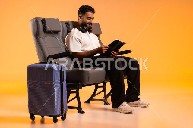 الجلوس في صالة المطار, التعلم والتثقف الذاتي، شغف التعرف على الكتب المحتلفة، الاستعداد للسفر في إجازة، بورتريه لرجل عربي خليجي سعودي يرتدي الزي الكاجوال يجلس على المقعد يمسك بيده كتاب وبجانبه حقيبة سفر, أهمية السياحة في السعودية, خلفية برتقالية