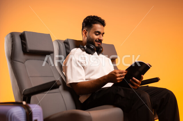 التعلم والتثقف الذاتي، شغف التعرف على الكتب المحتلفة، الاستعداد للسفر في إجازة، الجلوس في صالة المطار, بورتريه لرجل عربي خليجي سعودي يرتدي الزي الكاجوال يجلس على المقعد يمسك بيده كتاب وبجانبه حقيبة سفر, أهمية السياحة في السعودية, خلفية برتقالية