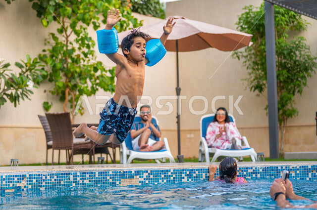 قضاء عطلة صيفية سعيدة، الأنشطة ترفيهية أثناء الإجازة، الاستعداد للقفز في المسبح، الاستعمتاع بالأجواء الصيفية العائلية، ولد عربي خليجي سعودي يرتدي عوامة السباحة ويقفز في بركة مائية