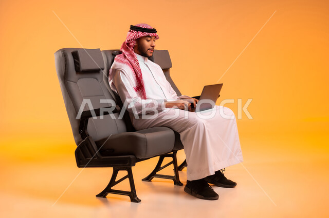 Sharing trip details on social media, a business trip, preparing to travel on vacation, sitting in the airport lounge, portrait of a Saudi Gulf Arab man wearing a traditional thobe and shemagh sitting on a seat using a laptop, the importance of tourism in Saudi Arabia, orange background