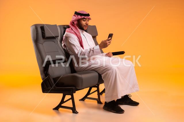 إجراء مكالمات الفيديو، أهمية السياحة في السعودية, استخدام الأجهزة التكنولوجية الحديثة، الاستعداد للسفر في إجازة، الجلوس في صالة المطار, بورتريه لرجل عربي خليجي سعودي يرتدي الثوب التقليدي والشماغ يجلس على المقعد يقوم باستخدام الهاتف المحمول, خلفية برتقالية