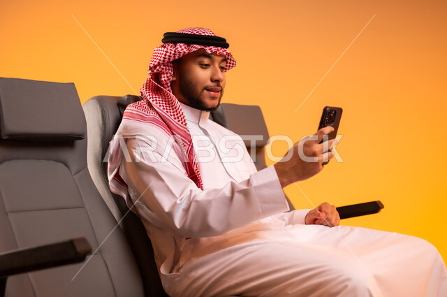 أهمية السياحة في السعودية, إجراء مكالمات فيديو،استخدام الأجهزة التكنولوجية الحديثة، الاستعداد للسفر في إجازة، الجلوس في صالة المطار, بورتريه لرجل عربي خليجي سعودي يرتدي الثوب التقليدي والشماغ يجلس على المقعد يقوم باستخدام الهاتف المحمول, خلفية برتقالية