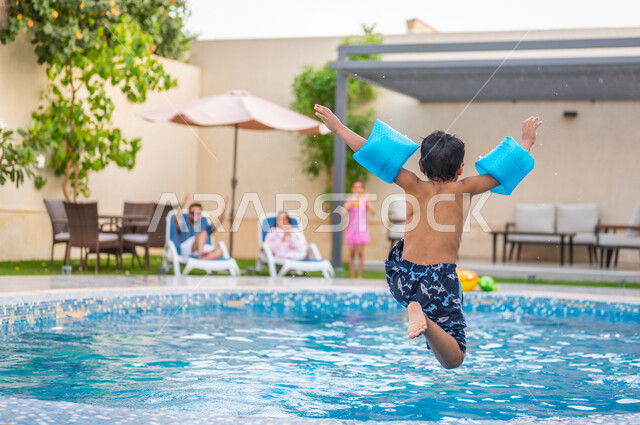 قضاء عطلة صيفية سعيدة، ارتداء عوامة السباحة للقفز في المسبح، رحلة سياحية ترفيهية، ولد عربي خليجي سعودي يقفز في بركة مائية، الاستمتاع بالأجواء الصيفية الممتعة