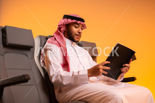 شغف التعرف على الكتب المحتلفة، قضاء وقت الفراغ بالتعلم والتثقف الذاتي، الاستعداد للسفر في إجازة، الجلوس في صالة المطار, بورتريه لرجل عربي خليجي سعودي يرتدي الثوب التقليدي والشماغ يجلس على المقعد يمسك بيده كتاب, أهمية السياحة في السعودية, خلفية برتقالية