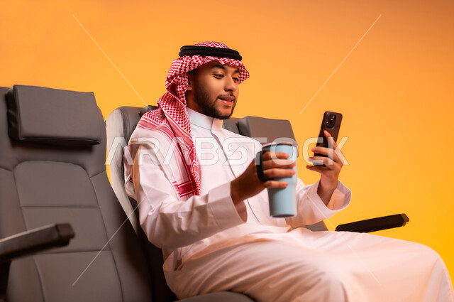 استخدام الأجهزة التقنية الحديثة والمتطورة، الاستمتاع بالمشروبات الساخنة، الاستعداد للسفر في إجازة صيفية، الجلوس في صالة المطار، أهمية السياحة في السعودية، بورتريه لشاب عربي خليجي سعودي يرتدي الثوب التقليدي يمسك بيده القهوة والهاتف المحمول، خلفية برتقالية