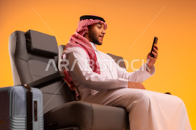 إجراء مكالمات فيديو، التواصل مع الأهل والأصدقاء، أهمية السياحة في السعودية, استخدام الهاتف المحمول، الاستعداد للسفر في إجازة، الجلوس في صالة المطار, بورتريه مقرب لرجل عربي خليجي سعودي يرتدي الثوب التقليدي يجلس على المقعد وبجانبه حقيبة سفر, خلفية برتقالية