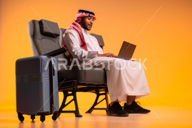 العمل عن بعد ، الجلوس في صالة المطار, بورتريه لرجل عربي خليجي سعودي يرتدي الثوب التقليدي والشماغ يجلس على المقعد يقوم باستخدام الحاسوب المحمول وبجانبه حقيبة سفر رمادية, أهمية السياحة في السعودية, مشاركة تفاصيل الرحلة على مواقع التواصل الاجتماعي، خلفية برتقالية