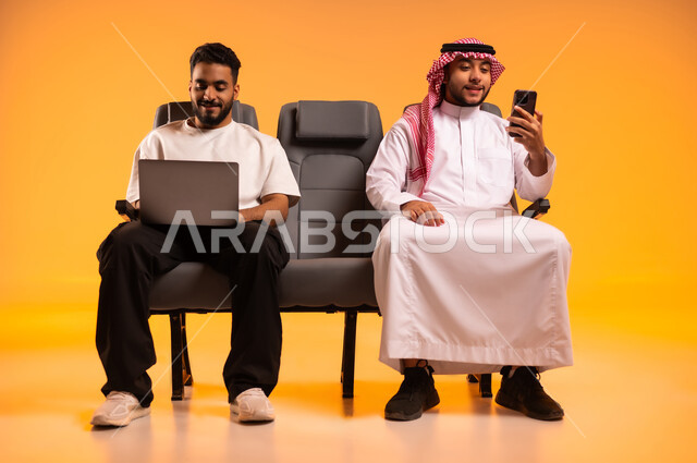 مشاركة تفاصيل الرحلة على مواقع التواصل الاجتماعي، استخدام الأجهزة التكنولوجية الحديثة، الاستعداد للسفر في إجازة، الجلوس في صالة المطار, بورتريه لرجل عربي خليجي سعودي يرتدي الزي الكاجوال يجلس على المقعد يقوم باستخدام الحاسوب المحمول يجلس مع صديقه, خلفية برتقالية
