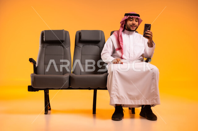 أهمية السياحة في السعودية, إجراء مكالمات فيديو،استخدام الأجهزة التكنولوجية الحديثة، الاستعداد للسفر في إجازة، الجلوس في صالة المطار, بورتريه لرجل عربي خليجي سعودي يرتدي الثوب التقليدي والشماغ يجلس على المقعد يقوم باستخدام الهاتف المحمول, خلفية برتقالية