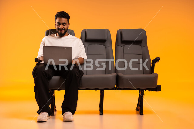 مشاركة تفاصيل الرحلة على مواقع التواصل الاجتماعي، الاستعداد للسفر في إجازة، الجلوس في صالة المطار, بورتريه لرجل عربي خليجي سعودي يرتدي الزي الكاجوال يجلس على المقعد يقوم باستخدام الحاسوب المحمول, أهمية السياحة في السعودية, رحلة عمل جوية، خلفية برتقالية