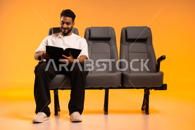 قضاء وقت الفراغ بالتعلم والتثقف الذاتي، شغف التعرف على الكتب المحتلفة، الاستعداد للسفر في إجازة، الجلوس في صالة المطار, بورتريه لرجل عربي خليجي سعودي يرتدي الزي الكاجوال يجلس على المقعد يمسك بيده كتاب, أهمية السياحة في السعودية, خلفية برتقالية
